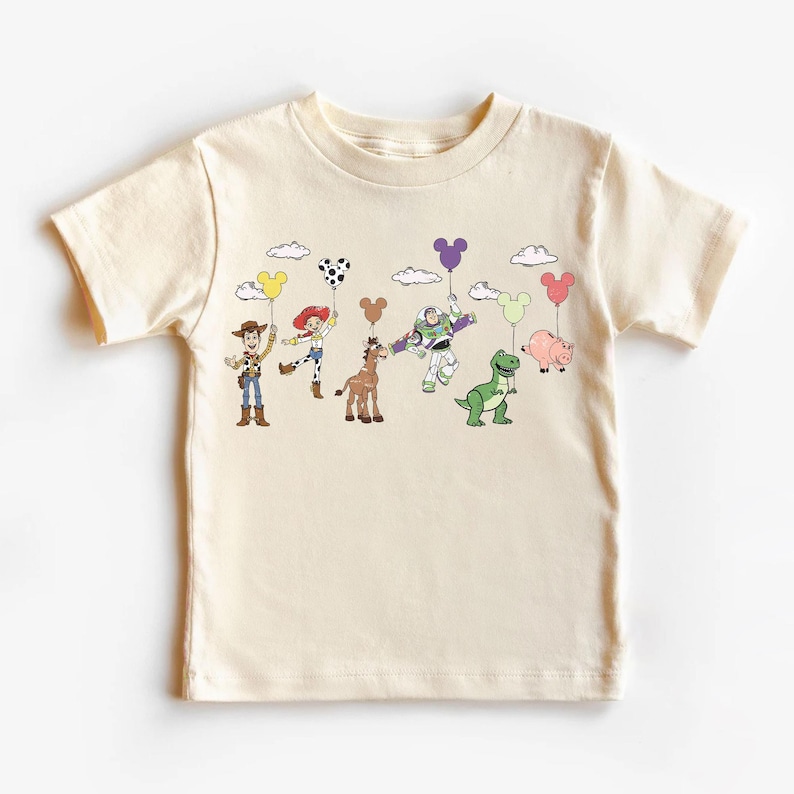 Op de afbeelding: Cr&egrave;mekleurig T-shirt met een kleurrijke afbeelding van stripfiguren die ballonvormige ballonnen vasthouden. De personages zijn onder meer een cowboy, cowgirl, paard, ruimteranger, dinosaurus en varken. De ballonnen zijn in verschillende kleuren en vormen.