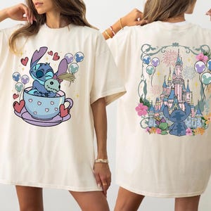 May include: An off-white t-shirt featuring a front graphic of a blue alien character sitting in a teacup with hearts and balloons. The back graphic displays the same blue alien character looking at a large castle with fireworks and balloons.