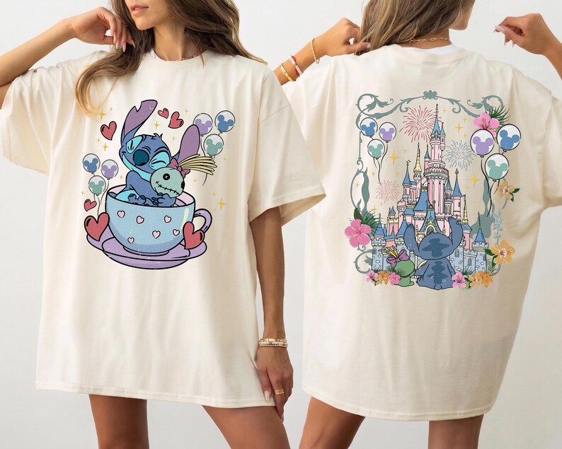 May include: An off-white t-shirt featuring a front graphic of a blue alien character sitting in a teacup with hearts and balloons. The back graphic displays the same blue alien character looking at a large castle with fireworks and balloons.