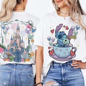 May include: Two white t-shirts featuring graphic prints. The back design shows a blue alien character and a doll in front of a castle with fireworks and balloons. The front design shows the same blue alien character sitting in a teacup with hearts.