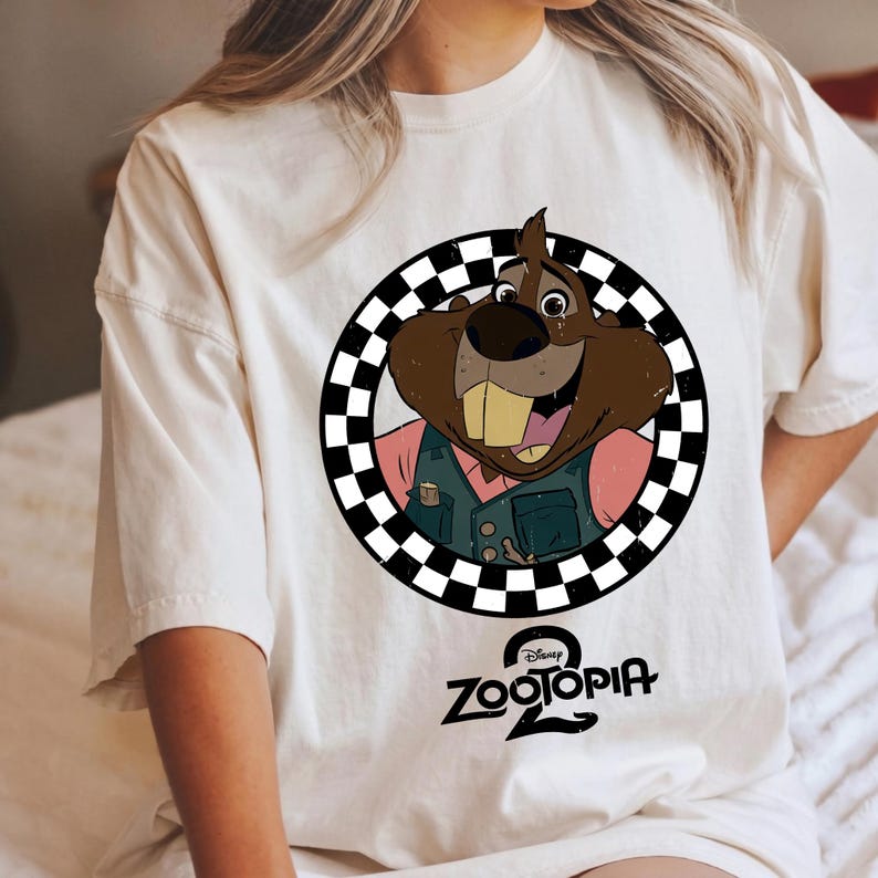 May include: An off-white t-shirt featuring a graphic of a brown beaver character from Zootopia 2 inside a black and white checkered circle. The beaver wears a teal vest over a pink shirt. The Zootopia 2 logo is printed in black below the circle.