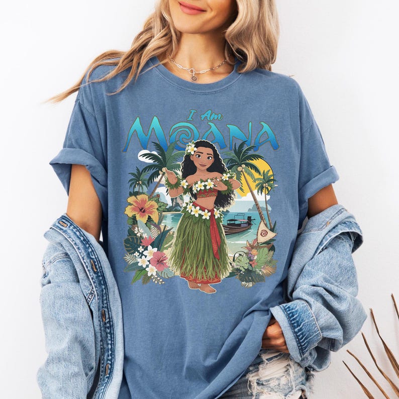 May include: Blue t-shirt featuring a colorful graphic of a woman in a grass skirt, floral lei, and the text "I Am Moana." The design includes palm trees, flowers, a boat, and a sunset. The shirt is made of a soft material.