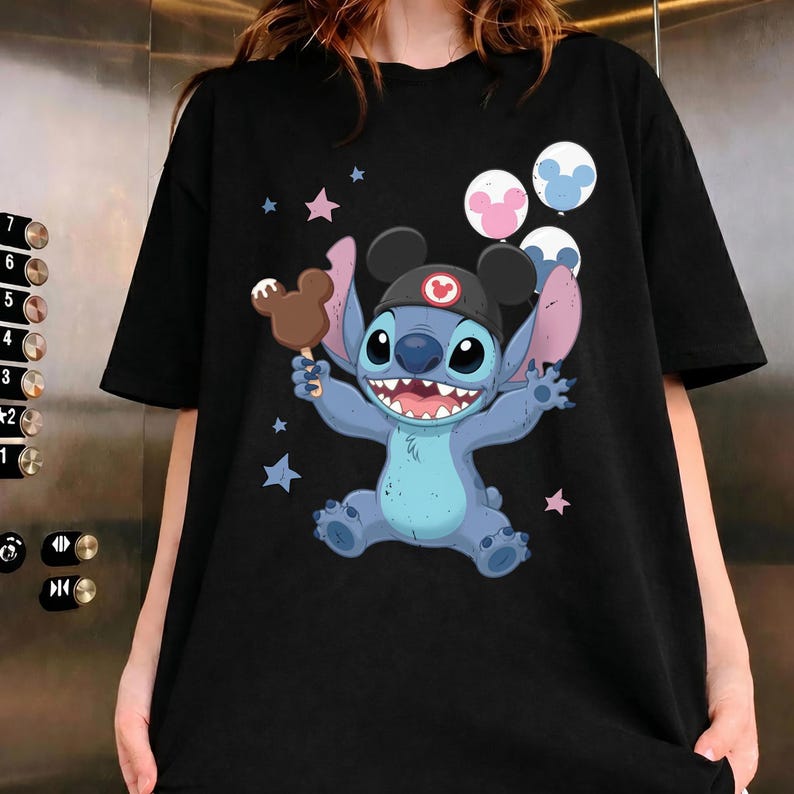 May include: A black short-sleeve t-shirt featuring a graphic of a blue alien character wearing a black cap with mouse ears, holding a brown chocolate-covered treat, surrounded by pink and blue stars and three round balloons with mouse ear shapes.
