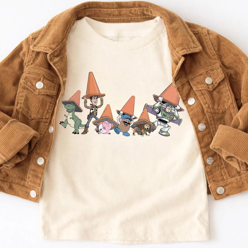 May include: Cream-colored t-shirt featuring a graphic of animated characters wearing orange traffic cones. A brown corduroy jacket is draped over the shirt. The characters include a dinosaur, a cowboy, a pig, a potato head, a dog, and Buzz Lightyear.