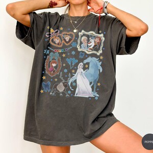 Puede incluir: Camiseta gris oscuro con un gr&aacute;fico colorido de personajes de la pel&iacute;cula animada Frozen. El dise&ntilde;o incluye im&aacute;genes de Elsa, Anna, Olaf y Sven, con detalles en azul y amarillo. La camiseta tiene un corte holgado.