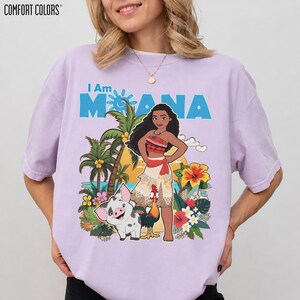 May include: A light purple Comfort Colors t-shirt featuring the text "I Am Moana" in blue. The graphic includes an illustration of Moana, a pig, a rooster, palm trees, and tropical flowers. The shirt is a relaxed fit.