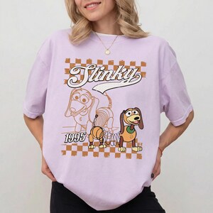 May include: A light purple t-shirt featuring a cartoon image of Slinky Dog from Toy Story. The design includes the word "Slinky" in a retro font, a speech bubble, and the year "1995". The background has a brown and white checkered pattern.