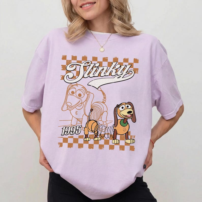 May include: A light purple t-shirt featuring a cartoon image of Slinky Dog from Toy Story. The design includes the word "Slinky" in a retro font, a speech bubble, and the year "1995". The background has a brown and white checkered pattern.
