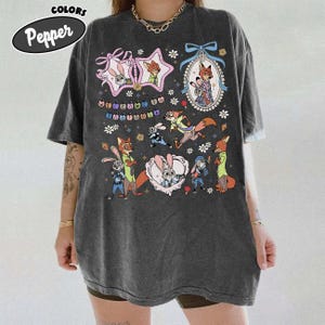 May include: A charcoal gray oversized t-shirt featuring a graphic print of characters from Zootopia, including Judy Hopps and Nick Wilde. The design includes floral accents, decorative frames, and the text Welcome to Zootopia in colorful letters.