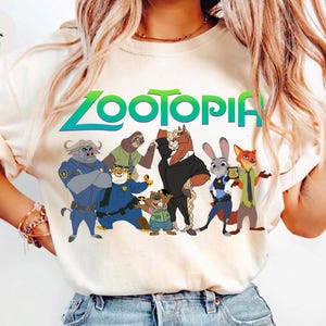 K&ouml;nnte beinhalten: Elfenbeinfarbenes T-Shirt mit dem Wort "Zootopia" in Gr&uuml;n und Blau sowie Zeichentrickfiguren aus dem Film. Zu den Figuren geh&ouml;ren ein B&uuml;ffel, ein Faultier, ein Pferd, ein Kaninchen und ein Fuchs. Das T-Shirt hat kurze &Auml;rmel.