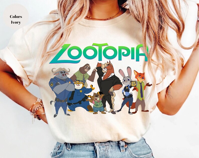 K&ouml;nnte beinhalten: Elfenbeinfarbenes T-Shirt mit dem Wort "Zootopia" in Gr&uuml;n und Blau sowie Zeichentrickfiguren aus dem Film. Zu den Figuren geh&ouml;ren ein B&uuml;ffel, ein Faultier, ein Pferd, ein Kaninchen und ein Fuchs. Das T-Shirt hat kurze &Auml;rmel.