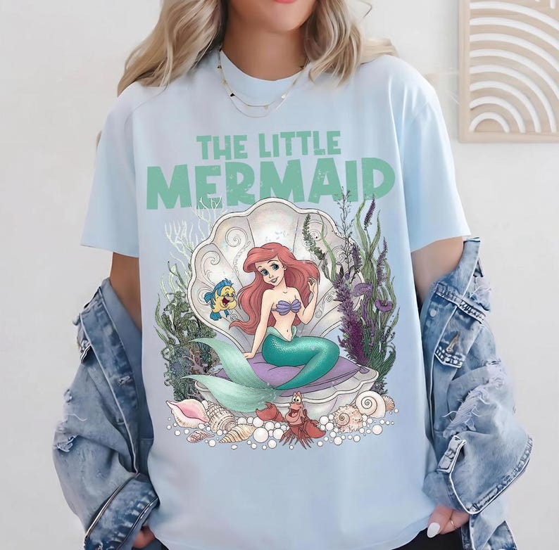 May include: Light blue t-shirt featuring the text "THE LITTLE MERMAID" in green. The graphic depicts Ariel, a mermaid with red hair and a teal tail, seated in a seashell. Flounder and Sebastian are also present in the design.