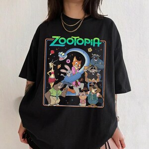 May include: A black short-sleeve t-shirt featuring a colorful graphic print of Zootopia characters including Nick Wilde, Judy Hopps, and Flash. The design includes the text Zootopia in a bright green font, surrounded by a thin orange border.