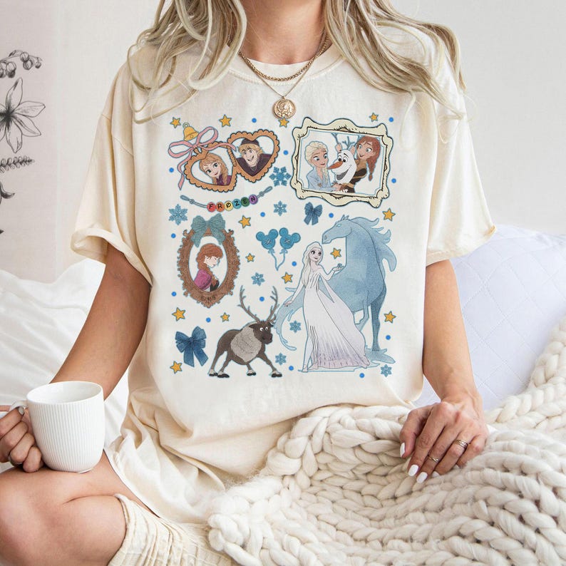 Puede incluir: Camiseta color crema con un gr&aacute;fico colorido de personajes de la pel&iacute;cula animada "Frozen". El dise&ntilde;o incluye retratos enmarcados, estrellas e im&aacute;genes de Elsa, Anna, Olaf y Sven. La palabra "Frozen" tambi&eacute;n es visible.
