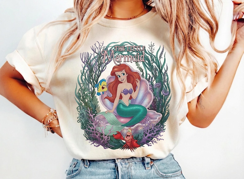 May include: Cream-colored t-shirt featuring a colorful graphic of Ariel from The Little Mermaid. The design includes Ariel, Flounder, Sebastian, and the words "The Little Mermaid" in a decorative font, surrounded by aquatic plants.