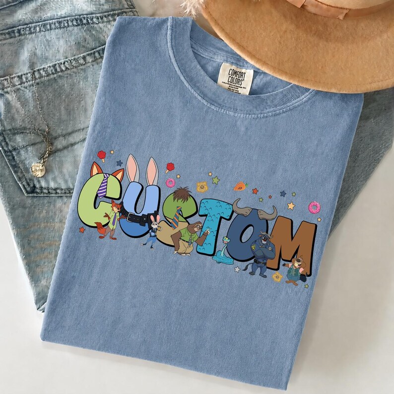 Pode incluir: Uma t-shirt azul com a palavra CUSTOM impressa em letras grandes e coloridas em estilo bolha. Cada letra apresenta uma personagem animal diferente do filme Zootopia, incluindo uma raposa, coelho, bicho-pregui&ccedil;a e touro, com pequenas estrelas.