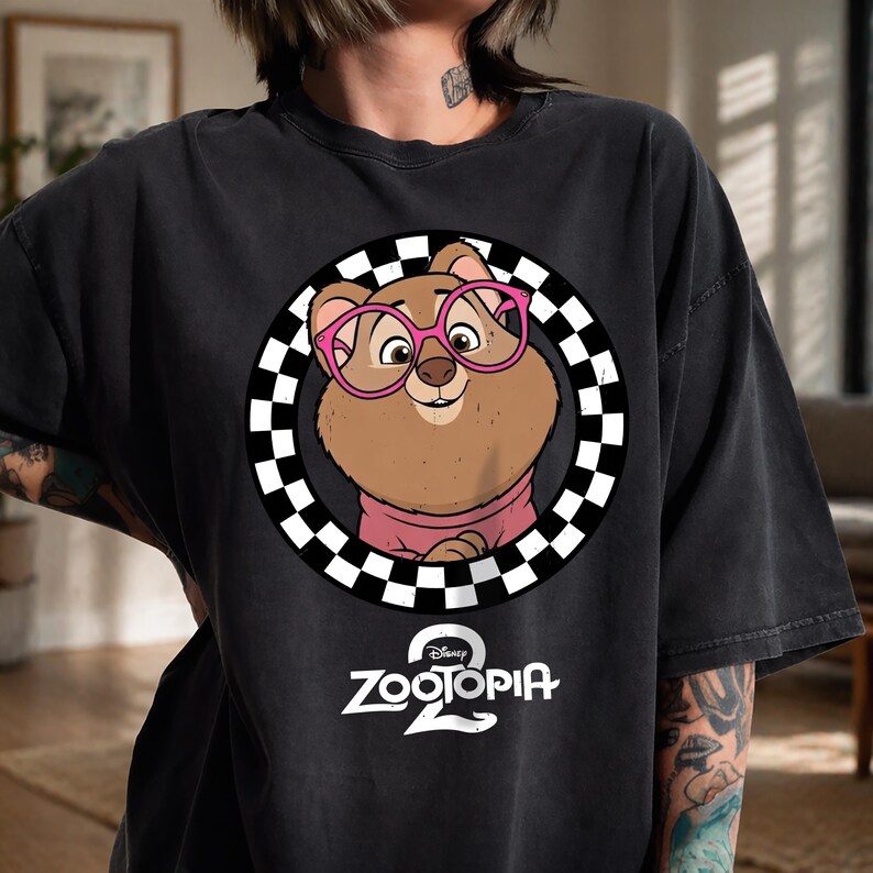 May include: A black short-sleeved t-shirt featuring a circular graphic of a brown animal wearing pink glasses inside a black and white checkered border. Below the graphic, the white text reads Disney Zootopia 2 in a stylized font.
