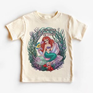 May include: Cream-colored t-shirt featuring a colorful graphic of Ariel from The Little Mermaid. Ariel is seated on a seashell, surrounded by aquatic plants, with Flounder and Sebastian. The text "The Little Mermaid" is arched above the image.