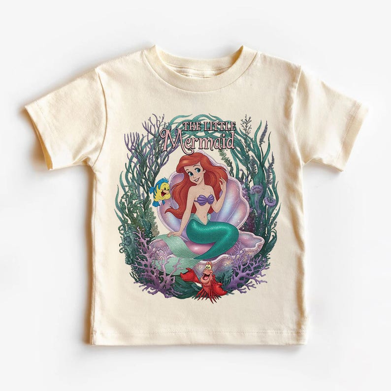 May include: Cream-colored t-shirt featuring a colorful graphic of Ariel from The Little Mermaid. Ariel is seated on a seashell, surrounded by aquatic plants, with Flounder and Sebastian. The text "The Little Mermaid" is arched above the image.