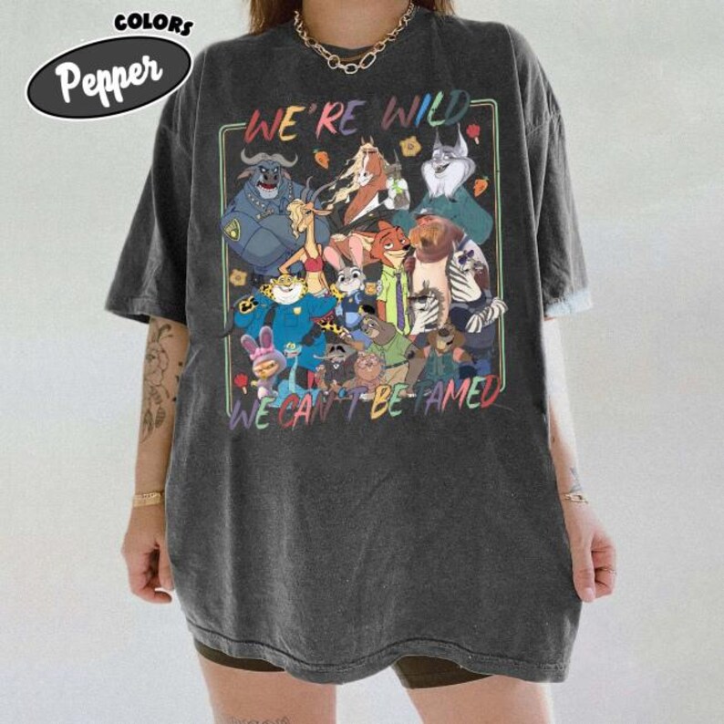 May include: A charcoal gray short-sleeve t-shirt featuring a colorful graphic print of various animated animal characters. The text "WE'RE WILD" appears at the top and "WE CAN'T BE TAMED" at the bottom of the rectangular graphic design.