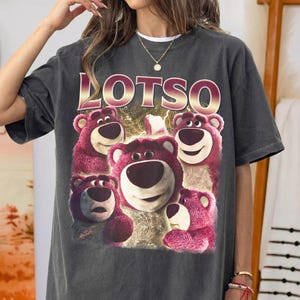 May include: Dark gray t-shirt featuring a graphic of Lotso, the pink bear from Toy Story, surrounded by other bears. The word "LOTSO" is printed in a gold and pink font above the bears. The shirt has a vintage, worn look.