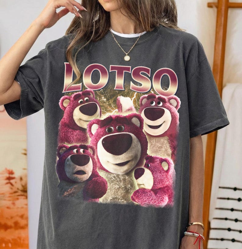 May include: Dark gray t-shirt featuring a graphic of Lotso, the pink bear from Toy Story, surrounded by other bears. The word "LOTSO" is printed in a gold and pink font above the bears. The shirt has a vintage, worn look.