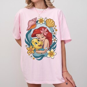 May include: A light pink t-shirt featuring a colorful graphic of a red-haired mermaid hugging a yellow fish. The design is framed by blue waves, seashells, and yellow flowers. The shirt has short sleeves and a relaxed fit.