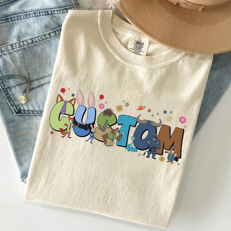 Pode incluir: Uma t-shirt de cor creme com a palavra CUSTOM em letras grandes e coloridas de estilo bloco. Cada letra incorpora v&aacute;rias personagens animais animadas e pequenos &iacute;cones como donuts, estrelas e cenouras espalhados pelo texto.