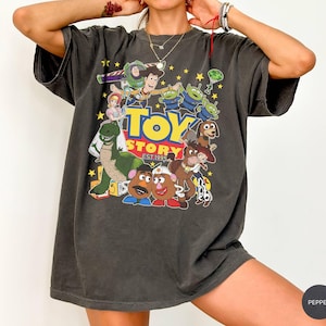 May include: Dark gray t-shirt featuring a colorful graphic of Toy Story characters, including Buzz Lightyear, Woody, and the Little Green Men. The text "Toy Story EST. 1995" is also visible. The shirt has a vintage, worn look.