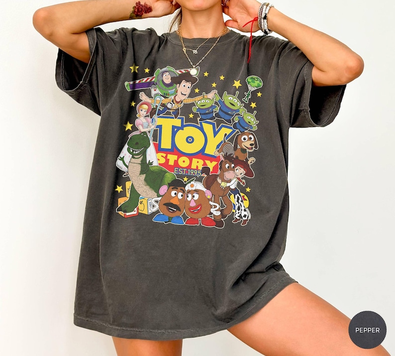 May include: Dark gray t-shirt featuring a colorful graphic of Toy Story characters, including Buzz Lightyear, Woody, and the Little Green Men. The text "Toy Story EST. 1995" is also visible. The shirt has a vintage, worn look.