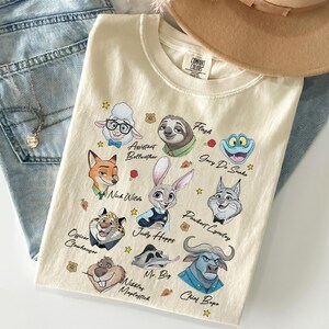 May include: A cream-colored short-sleeve t-shirt featuring a graphic print of various animated animal characters from Zootopia. The characters include Judy Hopps, Nick Wilde, and others, each with their name printed in black script text below them.