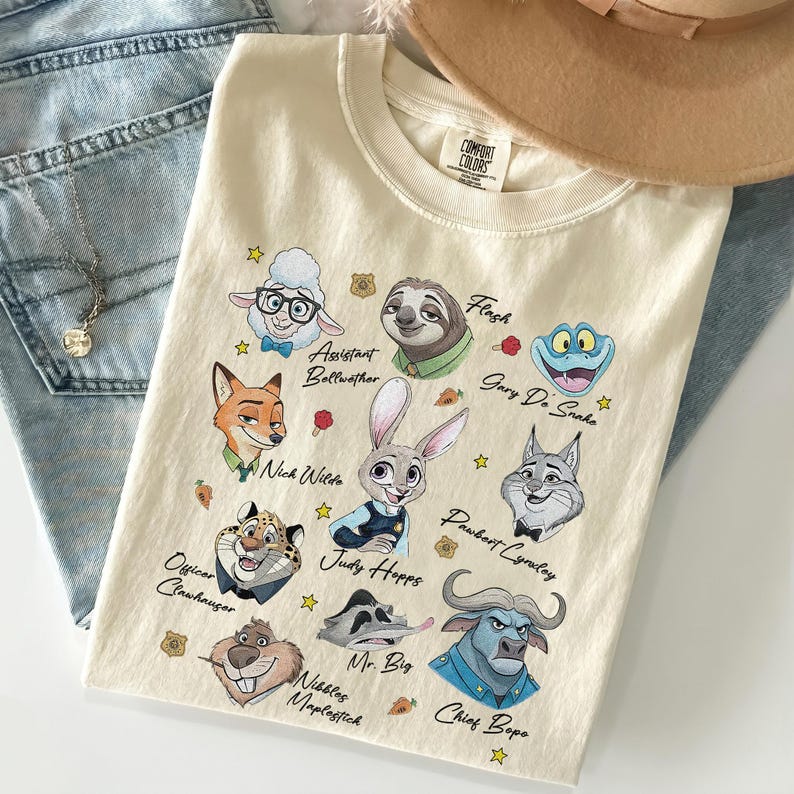May include: A cream-colored short-sleeve t-shirt featuring a graphic print of various animated animal characters from Zootopia. The characters include Judy Hopps, Nick Wilde, and others, each with their name printed in black script text below them.