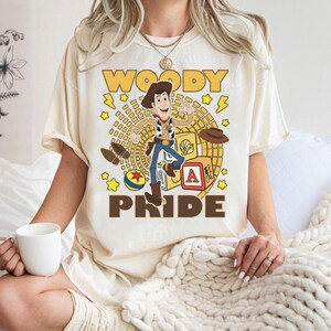 May include: Cream-colored t-shirt featuring a graphic of Woody from Toy Story. The design includes the words "WOODY PRIDE" in brown, along with cartoon illustrations of Woody, a hat, a block with the letter "A", and other elements.