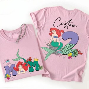 May include: Two light pink t-shirts with a mermaid theme. One shirt features the word "MOM" with a mermaid graphic, and the other has a mermaid with the number "2" and the word "Custom". Both shirts have a round neckline.