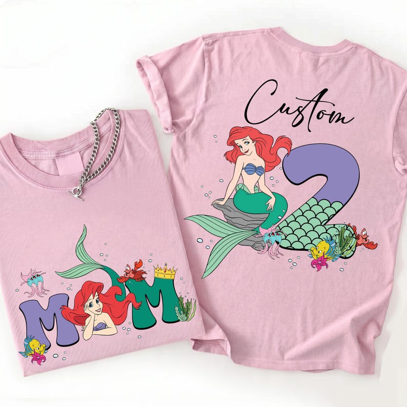 May include: Two light pink t-shirts with a mermaid theme. One shirt features the word "MOM" with a mermaid graphic, and the other has a mermaid with the number "2" and the word "Custom". Both shirts have a round neckline.