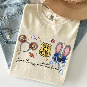 May include: A cream-colored t-shirt featuring a graphic print of Zootopia-themed icons including mouse ears, a police badge, a carrot, and a police hat with bunny ears. The text "Don't mess with the bunny" is printed in black cursive script below.