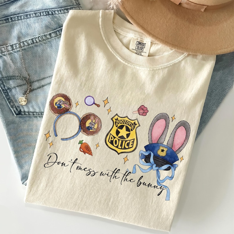 May include: A cream-colored t-shirt featuring a graphic print of Zootopia-themed icons including mouse ears, a police badge, a carrot, and a police hat with bunny ears. The text "Don't mess with the bunny" is printed in black cursive script below.