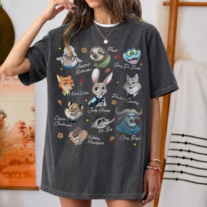 May include: A charcoal gray short-sleeve t-shirt featuring a graphic print of various animated characters from Zootopia, including Judy Hopps, Nick Wilde, and Chief Bogo, with their names printed in white script text alongside small star and carrot icons.