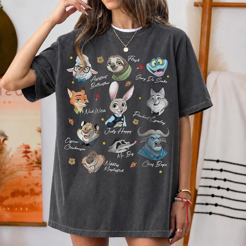 May include: A charcoal gray short-sleeve t-shirt featuring a graphic print of various animated characters from Zootopia, including Judy Hopps, Nick Wilde, and Chief Bogo, with their names printed in white script text alongside small star and carrot icons.