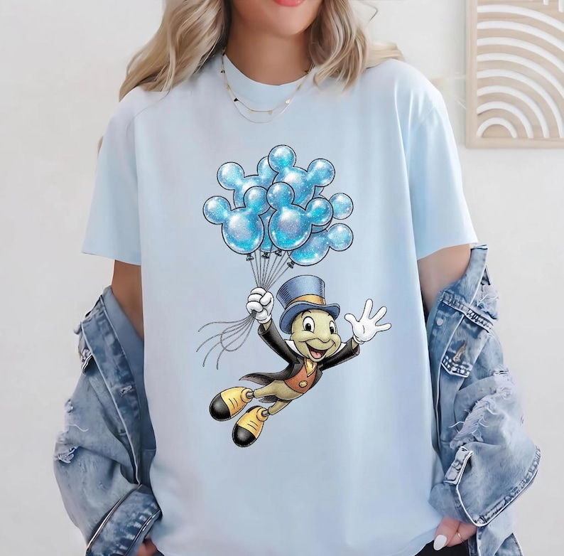 May include: Light blue t-shirt featuring a cartoon character holding a bunch of blue Mickey Mouse-shaped balloons. The character is wearing a top hat, black jacket, and yellow shoes. The balloons have a glittery effect.