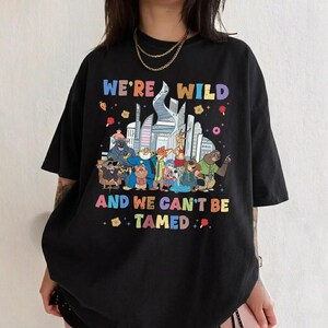 K&ouml;nnte beinhalten: Ein schwarzes T-Shirt mit kurzen &Auml;rmeln und einer bunten Grafik verschiedener animierter Tierfiguren vor einer Stadtsilhouette. Der Text WE'RE WILD AND WE CAN'T BE TAMED steht in mehrfarbigen Buchstaben &uuml;ber und unter den Charakteren.
