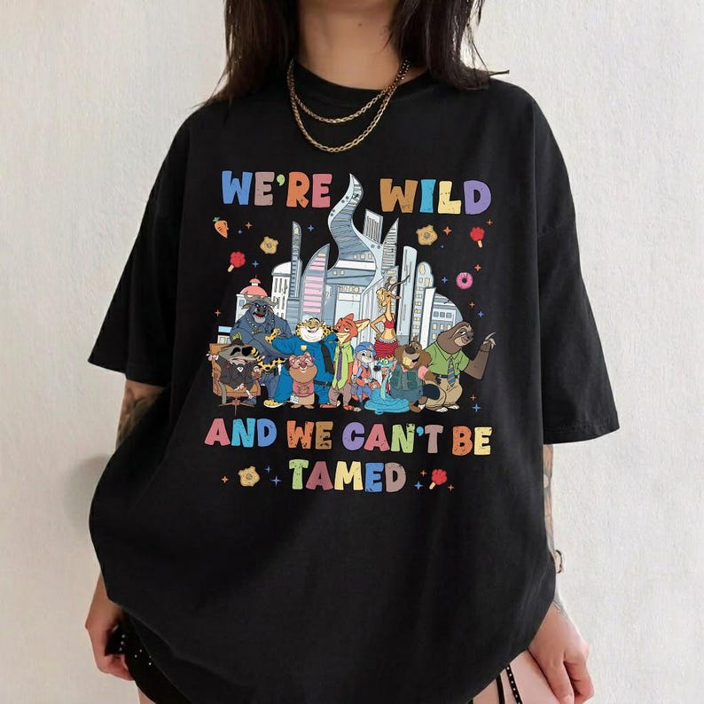 K&ouml;nnte beinhalten: Ein schwarzes T-Shirt mit kurzen &Auml;rmeln und einer bunten Grafik verschiedener animierter Tierfiguren vor einer Stadtsilhouette. Der Text WE'RE WILD AND WE CAN'T BE TAMED steht in mehrfarbigen Buchstaben &uuml;ber und unter den Charakteren.