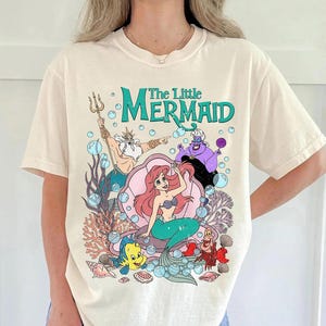 Puede incluir: Camiseta color crema con un gr&aacute;fico colorido de los personajes de "La Sirenita" de Disney. El dise&ntilde;o incluye a Ariel, el Rey Trit&oacute;n, &Uacute;rsula, Flounder y Sebasti&aacute;n, con el t&iacute;tulo en verde azulado. La obra de arte est&aacute; rodeada de burbujas y coral.
