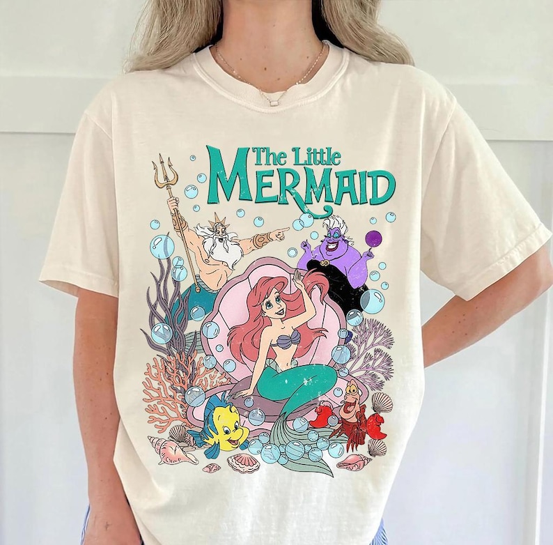 Puede incluir: Camiseta color crema con un gr&aacute;fico colorido de los personajes de "La Sirenita" de Disney. El dise&ntilde;o incluye a Ariel, el Rey Trit&oacute;n, &Uacute;rsula, Flounder y Sebasti&aacute;n, con el t&iacute;tulo en verde azulado. La obra de arte est&aacute; rodeada de burbujas y coral.