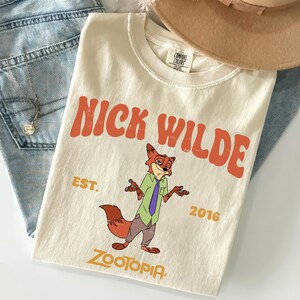 Pode incluir: Uma t-shirt de manga curta na cor creme com um gr&aacute;fico da personagem Nick Wilde de Zootopia. O texto diz NICK WILDE, EST. 2016 e ZOOTOPIA em letras laranja e amarelas. A t-shirt tem gola redonda e uma etiqueta da marca Comfort Colors.