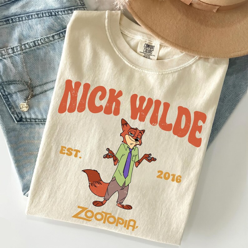 Pode incluir: Uma t-shirt de manga curta na cor creme com um gr&aacute;fico da personagem Nick Wilde de Zootopia. O texto diz NICK WILDE, EST. 2016 e ZOOTOPIA em letras laranja e amarelas. A t-shirt tem gola redonda e uma etiqueta da marca Comfort Colors.