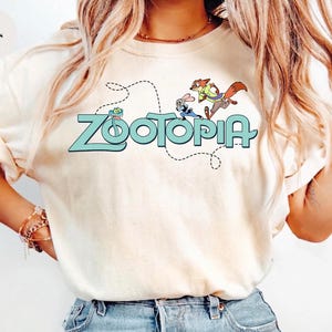 May include: An ivory t-shirt featuring a graphic print of the word Zootopia in light blue letters. Above the text, a small green snake, a rabbit, and a fox are illustrated. A dashed black line curves around the characters and the text.