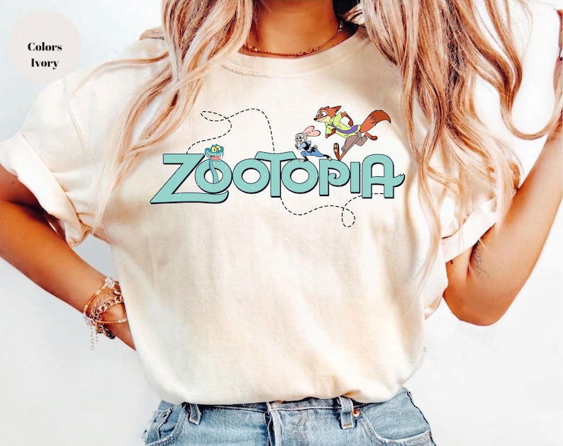 May include: An ivory t-shirt featuring a graphic print of the word Zootopia in light blue letters. Above the text, a small green snake, a rabbit, and a fox are illustrated. A dashed black line curves around the characters and the text.