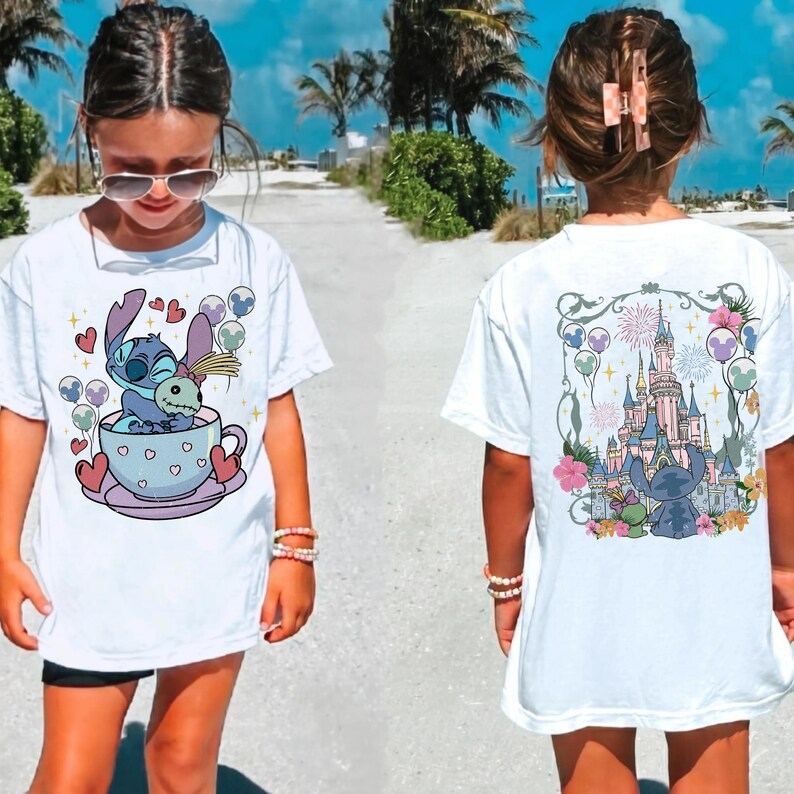 May include: A white short-sleeve t-shirt featuring a graphic of Stitch sitting in a teacup on the front and a graphic of Stitch standing before a castle with fireworks and floral accents on the back. The design includes hearts and balloons.