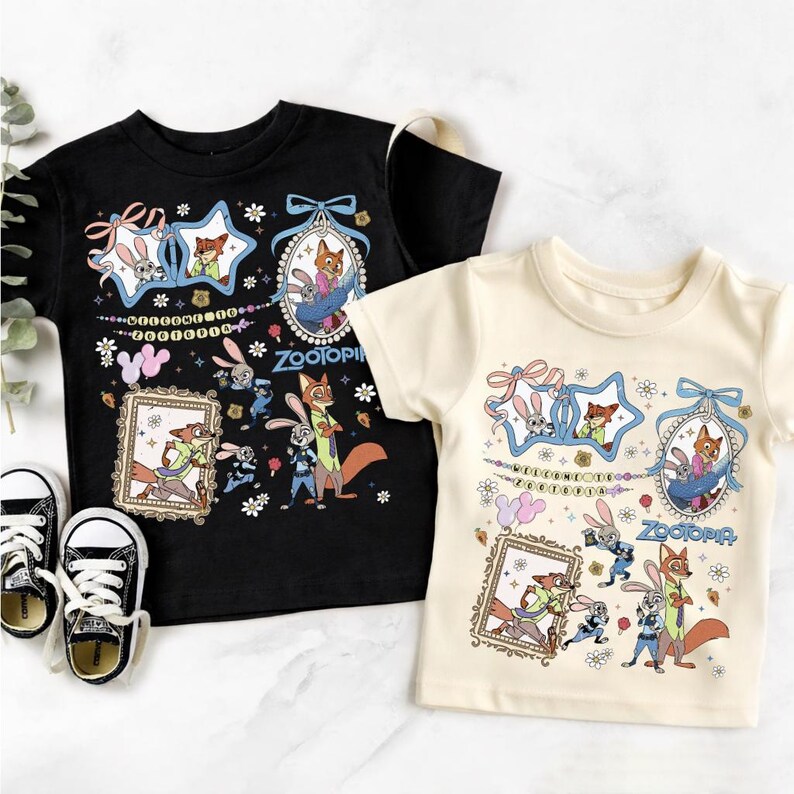 May include: A black t-shirt and a cream t-shirt, each featuring a Zootopia graphic print with characters Judy Hopps and Nick Wilde, floral accents, framed portraits, and the text Welcome to Zootopia, alongside a pair of black and white sneakers.